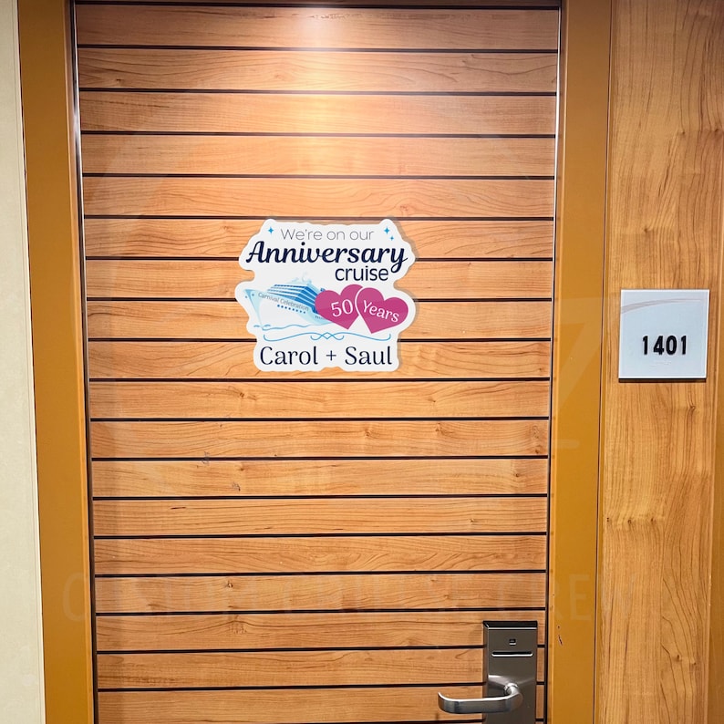 Op de afbeelding: Een houten deur met een sticker waarop staat "We're on our Anniversary cruise 50 Years Carol + Saul". De deur heeft het nummer "1401" aan de zijkant.