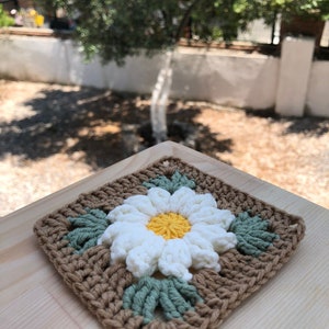 May include: A crocheted square with a white daisy in the center. The daisy has a yellow center and green leaves. The square is brown with green leaves around the daisy.