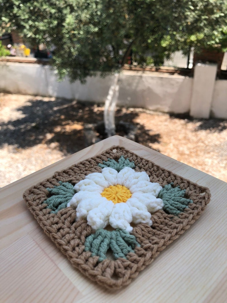 May include: A crocheted square with a white daisy in the center. The daisy has a yellow center and green leaves. The square is brown with green leaves around the daisy.