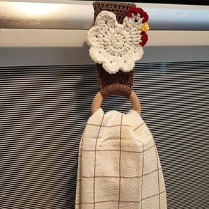 Op de afbeelding: Een handgemaakte gehaakte keukendoekhouder met een wit kippenontwerp met rode en gele accenten. De houder is bruin en heeft een houten ring om een geruite keukendoek vast te houden.