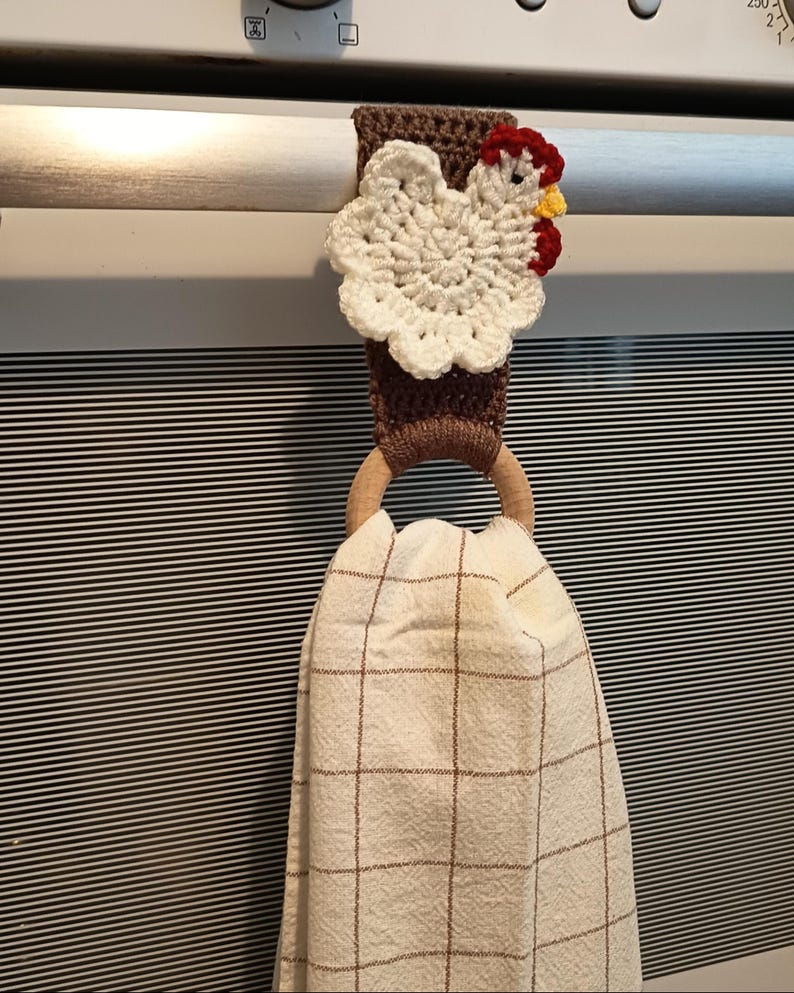 Op de afbeelding: Een handgemaakte gehaakte keukendoekhouder met een wit kippenontwerp met rode en gele accenten. De houder is bruin en heeft een houten ring om een geruite keukendoek vast te houden.