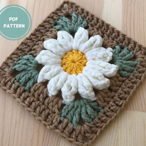 May include: A crochet square featuring a white daisy with a yellow center, surrounded by green leaves. The square is made with brown yarn.