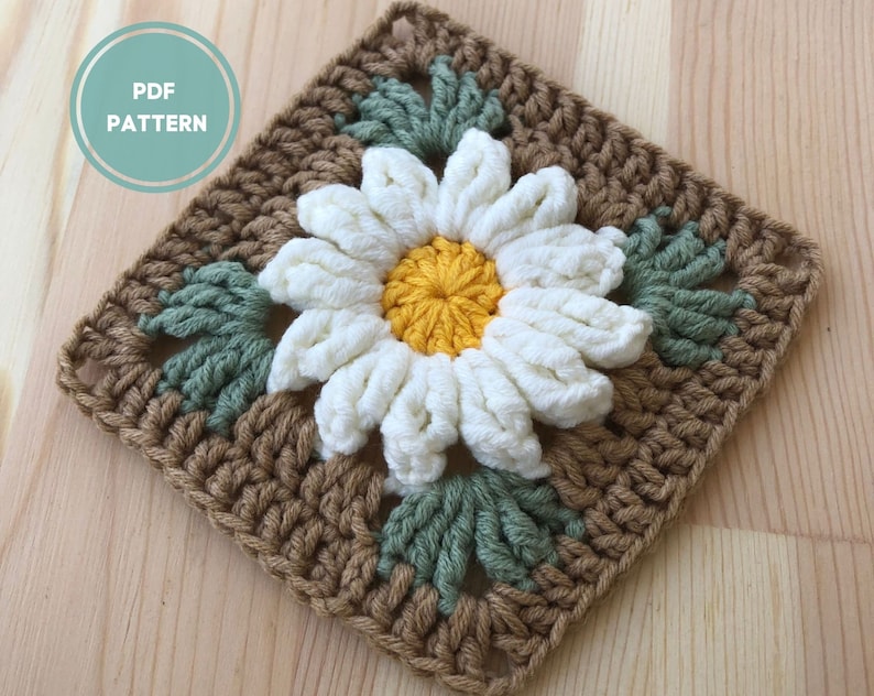 May include: A crochet square featuring a white daisy with a yellow center, surrounded by green leaves. The square is made with brown yarn.