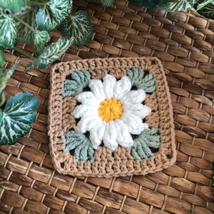 May include: A crocheted square with a white daisy flower in the center. The flower has a yellow center and green leaves around it. The square is made with brown and green yarn.