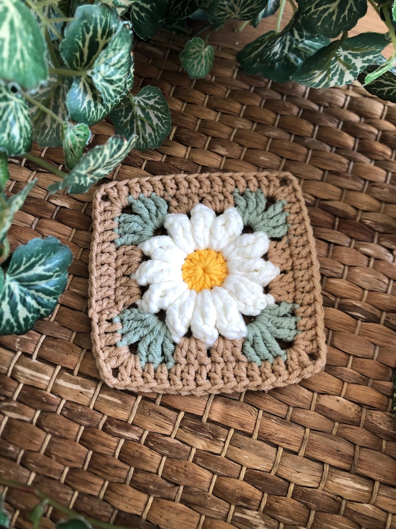 May include: A crocheted square with a white daisy flower in the center. The flower has a yellow center and green leaves around it. The square is made with brown and green yarn.