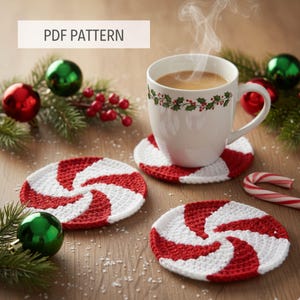 May include: A white mug with a holly berry design sits on a red and white crocheted coaster. Two additional coasters with a peppermint swirl design are visible. The image includes Christmas ornaments, greenery, and the text "PDF PATTERN".