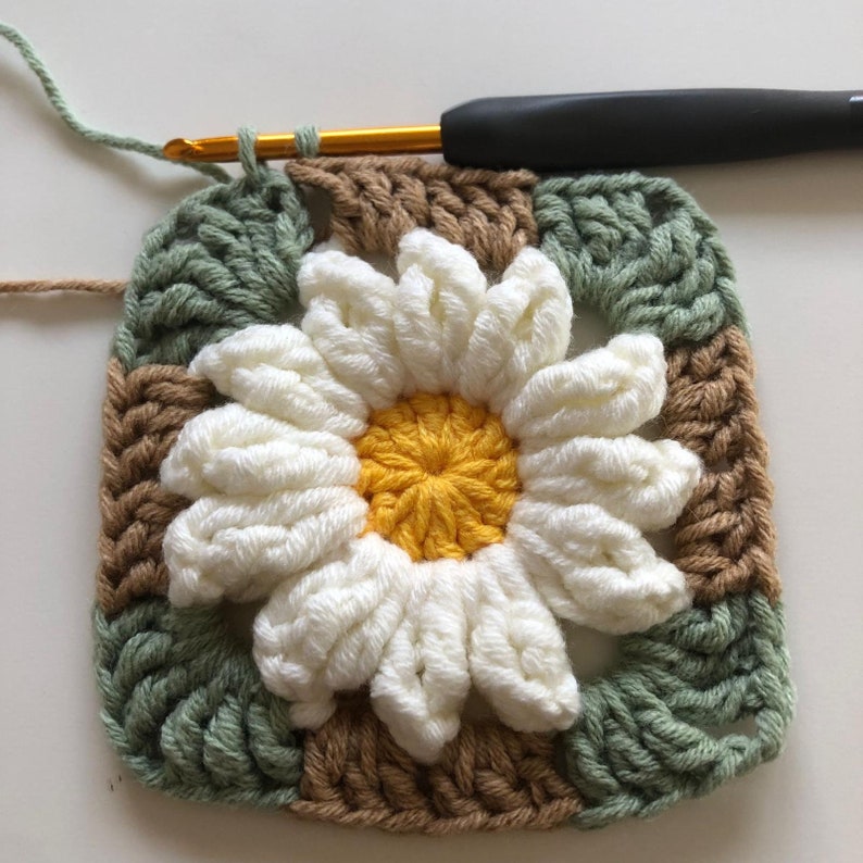 May include: A crocheted square with a white daisy flower in the center. The flower has a yellow center and is surrounded by brown and green squares.