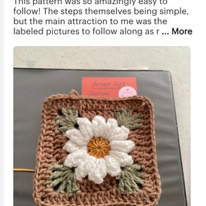 May include: A crocheted square with a brown background and a white daisy in the center. The daisy has a yellow center and green leaves around it. The square is surrounded by a brown border.