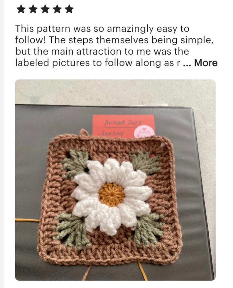 May include: A crocheted square with a brown background and a white daisy in the center. The daisy has a yellow center and green leaves around it. The square is surrounded by a brown border.