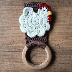 Op de afbeelding: Een handgemaakte gehaakte keukendoekhouder met een wit kippenontwerp met rode en gele accenten. De houder is bevestigd aan een bruine gehaakte band en een houten ring. De achtergrond is een houten oppervlak.