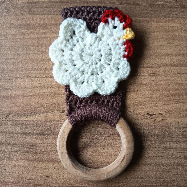 Op de afbeelding: Een handgemaakte gehaakte keukendoekhouder met een wit kippenontwerp met rode en gele accenten. De houder is bevestigd aan een bruine gehaakte band en een houten ring. De achtergrond is een houten oppervlak.
