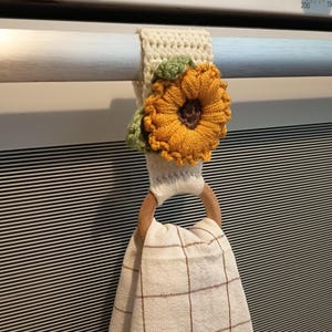 Op de afbeelding: Een cr&egrave;mekleurige, gehaakte ovenhanddoekhouder met een zonnebloemontwerp. De zonnebloem heeft gele bloemblaadjes, een bruin hart en groene bladeren. Een houten ring houdt een beige en bruin geruite keukendoek vast.