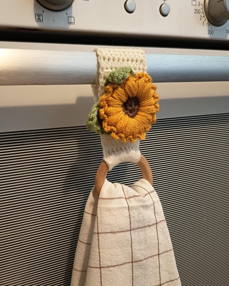 Op de afbeelding: Een cr&egrave;mekleurige, gehaakte ovenhanddoekhouder met een zonnebloemontwerp. De zonnebloem heeft gele bloemblaadjes, een bruin hart en groene bladeren. Een houten ring houdt een beige en bruin geruite keukendoek vast.