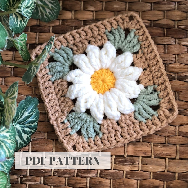 May include: A crochet square featuring a white daisy with a yellow center, surrounded by green leaves. The square is made with brown and green yarn. The text "PDF PATTERN" is visible at the bottom of the image.