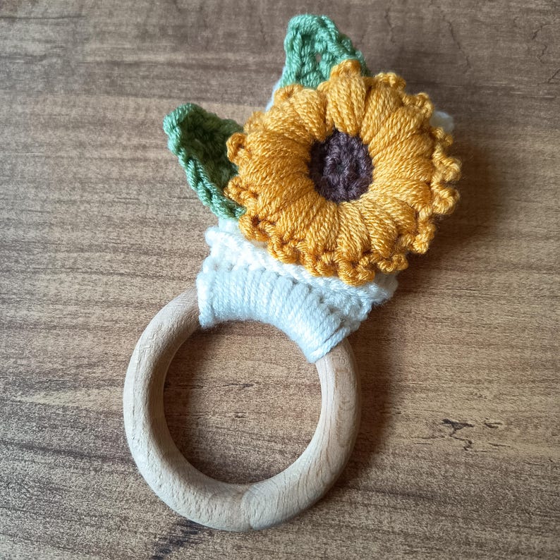 Op de afbeelding: Een handgemaakte houten bijtring voor baby's met een gehaakt zonnebloemontwerp. De zonnebloem heeft gele bloemblaadjes, een bruin hart en groene bladeren. De bijtring is gemaakt van lichtgekleurd hout.
