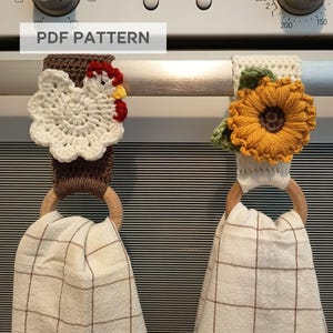 Op de afbeelding: Twee gehaakte keukendoekhouders met houten ringen. E&eacute;n heeft een witte kip met rode accenten, de andere een gele zonnebloem met groene bladeren. De handdoeken zijn cr&egrave;mekleurig met een bruin ruitpatroon. Tekst bovenaan luidt "PDF PATTERN".