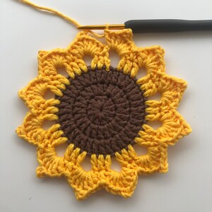 May include: A crocheted sunflower with a brown center and yellow petals. The sunflower is being worked on with a crochet hook.