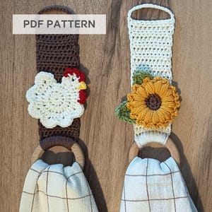 Op de afbeelding: Twee handgemaakte gehaakte handdoekhouders. E&eacute;n heeft een witte kip met rode en gele accenten, de andere een gele zonnebloem met groene bladeren. Beide hebben houten ringen en geruite handdoeken. De tekst "PDF PATTERN" is zichtbaar.