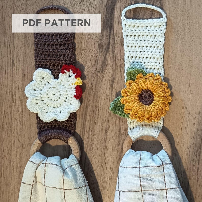 Op de afbeelding: Twee handgemaakte gehaakte handdoekhouders. E&eacute;n heeft een witte kip met rode en gele accenten, de andere een gele zonnebloem met groene bladeren. Beide hebben houten ringen en geruite handdoeken. De tekst "PDF PATTERN" is zichtbaar.