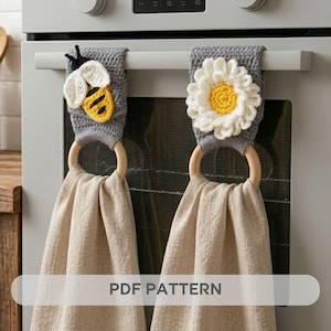 May include: Two gray crochet kitchen towel holders with wooden rings, one featuring a bee and the other a daisy. The towels are beige and hang from the oven handle. The text "PDF PATTERN" is visible.