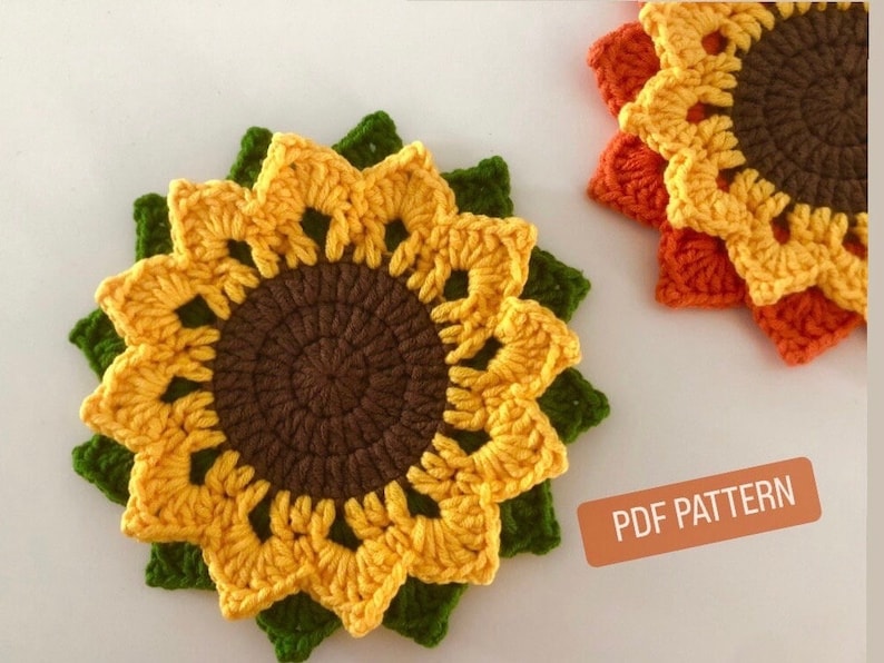 May include: Two crocheted sunflower coasters, one yellow and green, the other orange and brown. The coasters are made with a detailed crochet pattern and have a round center. The image includes the text "PDF PATTERN".