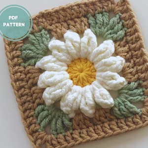May include: Crochet square with a white daisy flower in the center, surrounded by green leaves and a brown border.