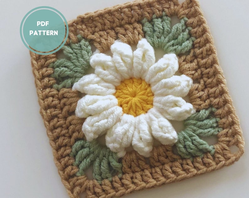 May include: Crochet square with a white daisy flower in the center, surrounded by green leaves and a brown border.