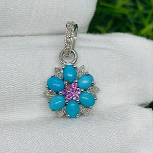 May include: A silver pendant featuring a turquoise and pink flower design with a white background. The pendant is made of silver and has a delicate design.