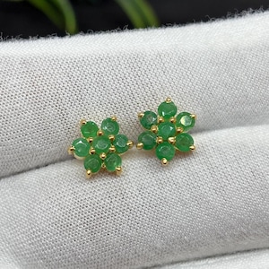 Puede incluir: Un par de pendientes de botón dorados con forma de flor. Cada pendiente presenta seis gemas verdes redondas y brillantes dispuestas en un patrón floral. Los pendientes se muestran sobre una superficie blanca texturizada.