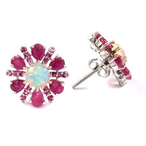 Puede incluir: Un par de pendientes de botón con forma de flor. Cada pendiente presenta una piedra central, redonda y opalescente, rodeada de gemas rosas en forma de lágrima. Los pendientes están engarzados en un metal plateado.