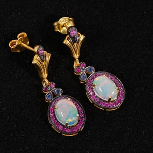 Puede incluir: Un par de pendientes dorados con ópalos ovales rodeados de pequeñas piedras rosas y azules. Los pendientes tienen un diseño decorativo y un cierre de poste.