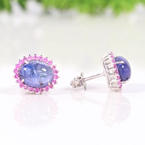 Puede incluir: Un par de pendientes de botón con piedras preciosas azules ovaladas rodeadas de pequeñas piedras preciosas rosas. Los pendientes tienen engastes y postes de metal plateado. El fondo está borroso con tonos verdes y blancos.