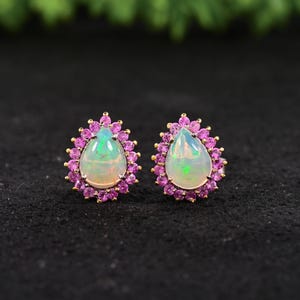Könnte beinhalten: Ein Paar goldene Ohrstecker mit tropfenförmigen Opalen, die jeweils von kleinen rosa Edelsteinen umgeben sind. Die Opale weisen einen milchig-weißen Farbton mit grünen und blauen Lichtreflexen auf. Die Ohrringe sind vor einem schwarzen Hintergrund platziert.