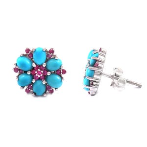 May include: A pair of flower-shaped stud earrings. Each earring features six oval turquoise stones surrounding a cluster of small, round, red gemstones. The earrings are set in a silver metal.