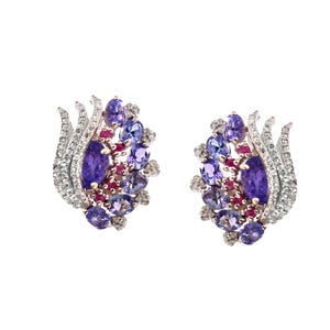 May include: A pair of ornate stud earrings. Each earring features a cluster of purple and violet gemstones, including oval and round cuts, accented with small red stones and clear crystals. The earrings have a silver-colored metal setting.