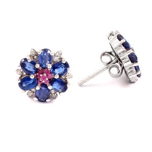 May include: A pair of flower-shaped stud earrings. Each earring features a cluster of blue oval gemstones surrounding a center of pink gemstones, with small white gemstones around the edges. The earrings are set in a silver metal.
