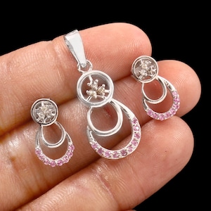 May include: A silver pendant and earring set. The pendant has a circular top with a floral design and a double-loop design with pink gemstones. The earrings have a similar design. The jewellery is set against a black background.