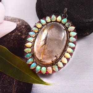 Puede incluir: Un colgante de plata con un cristal de cuarzo transparente con inclusiones doradas, rodeado por un halo de ópalos blancos y rubíes rojos.