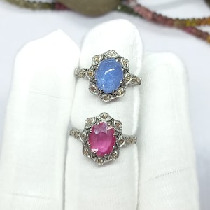 May include: Two silver rings with oval-shaped gemstones. The top ring has a light blue gemstone surrounded by smaller diamonds. The bottom ring has a pink gemstone surrounded by smaller diamonds.