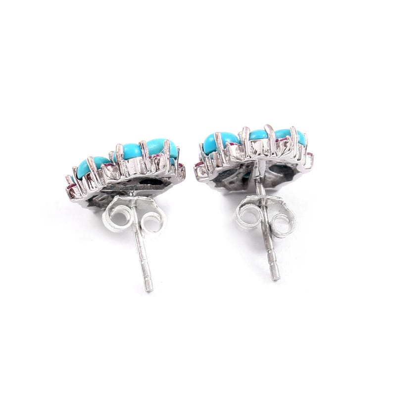 May include: A pair of silver-toned stud earrings. Each earring features a floral design with bright turquoise stones and small red accents. The earrings are set against a white background.
