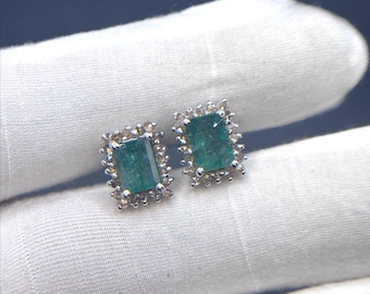 May include: A pair of emerald-cut emerald stud earrings. Each earring features a rectangular, deep green emerald stone surrounded by a halo of small, sparkling stones. The earrings are set in a silver-colored metal, creating a classic and elegant look.