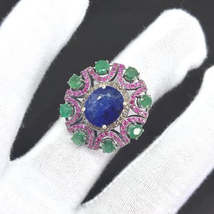 May include: An elaborate ring with a central oval blue gemstone, encircled by smaller green and pink stones. The ring has a silver band and a detailed, floral design. The gemstones are arranged in a circular pattern, creating a striking visual.