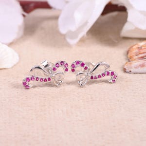 Puede incluir: Un par de pendientes de botón plateados con forma de lazo. Cada pendiente presenta un diseño de lazo con una línea de pequeñas piedras preciosas de color rosa brillante. Los pendientes están sobre un fondo neutro, resaltando su delicado diseño.