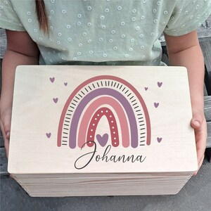 Könnte beinhalten: Eine Holzkiste mit einem Regenbogen-Design in Rosa, Lila und Rot und dem Namen "Johanna" in Kursivschrift. Die Kiste ist mit kleinen rosa Herzen verziert.