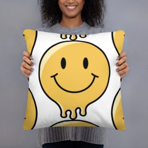 May include: White pillow with a yellow smiley face design. The smiley face is dripping with yellow liquid.