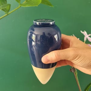 Op de afbeelding: Een marineblauwe keramische urn met een crèmekleurige basis en een bijpassend deksel. De urn wordt in een hand gehouden tegen een groene achtergrond. Het deksel heeft een decoratief ontwerp.