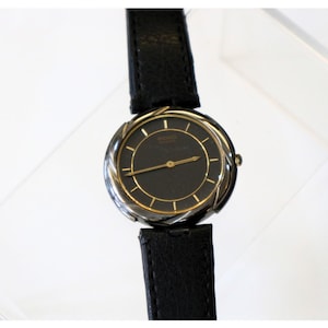 May include: A black wristwatch with a dark face and gold-coloured accents. The watch has a black leather strap. The watch face has gold-coloured hands and markers. The watch is a classic style.