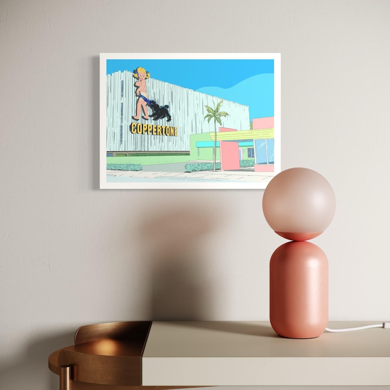 May include: A framed print featuring a retro illustration of a Coppertone advertisement with a woman and dog. The artwork is set against a blue sky and a building with a palm tree. A peach-colored lamp with a spherical shade sits on a neutral-toned surface.