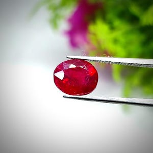 Puede incluir: Una piedra preciosa ovalada de color rojo oscuro sostenida por unas pinzas. La piedra preciosa está facetada y tiene un alto pulido.