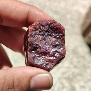 Könnte beinhalten: Ein roher, ungeschliffener Rubin-Edelstein mit tiefroter und dunkelvioletter Färbung. Der Stein weist eine hexagonale, kristalline Struktur mit einer rauen, texturierten Oberfläche auf, die natürliche Mineraleinschlüsse zeigt.